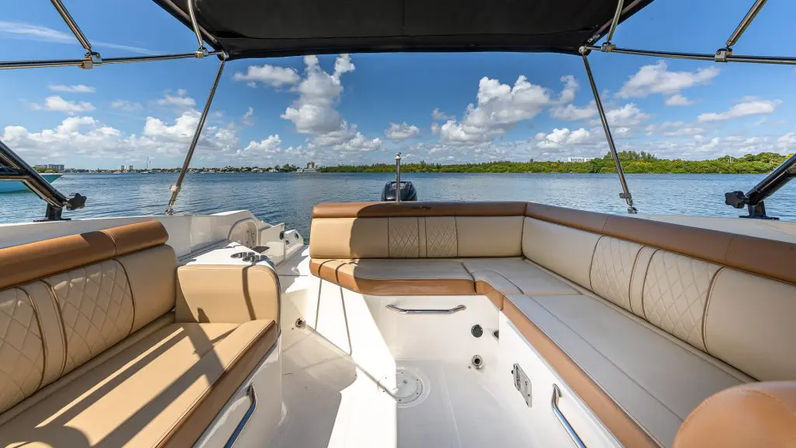 Sunlit motorboat deck with tan leather wraparound seating under a black bimini, facing calm blue coastal waters, puffy clouds and a distant shoreline.