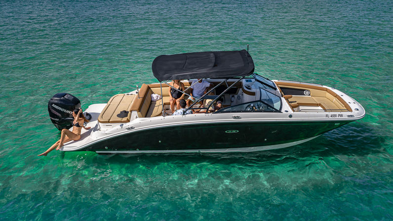 Aerial view of a sleek black-and-white motorboat with tan seating and a black bimini top anchored in clear turquoise water, friends relaxing onboard and a woman sunbathing on the stern.