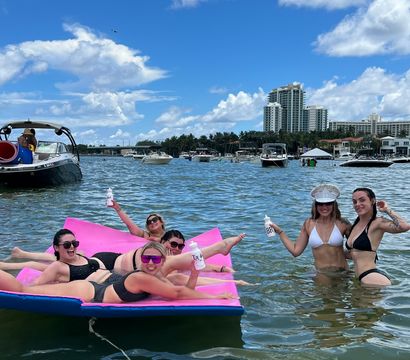 Women in bikinis lounging on a bright pink floating mat and wading in shallow water near anchored boats and yachts, with waterfront high-rise condos under a sunny blue sky with fluffy clouds.