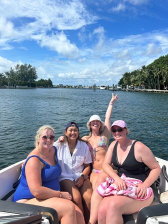 Four friends enjoying a sunny boat ride in a palm-lined coastal inlet, smiling in swimsuits under blue skies and fluffy clouds.