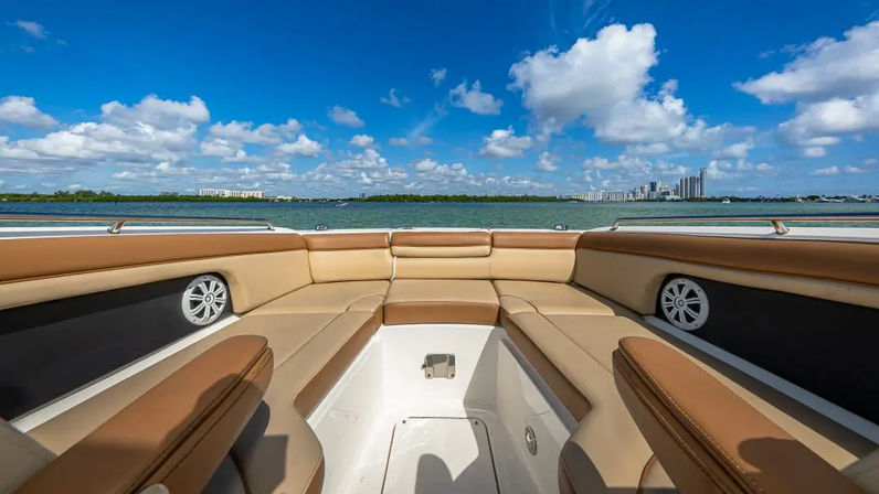 Sunlit tan leather bow seating on a luxury boat, looking across a blue bay toward a distant waterfront city skyline under fluffy white clouds.