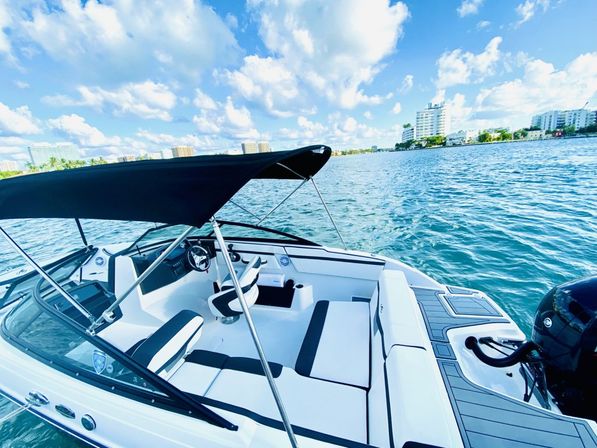 White and black motorboat with cushioned seating, steering helm and black bimini top on blue water near an urban waterfront with mid-rise buildings under a bright, partly cloudy sky.