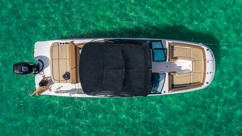 Aerial view of a sleek motorboat with tan seating and black canopy floating on crystal-clear turquoise water, a person lounging at the stern.