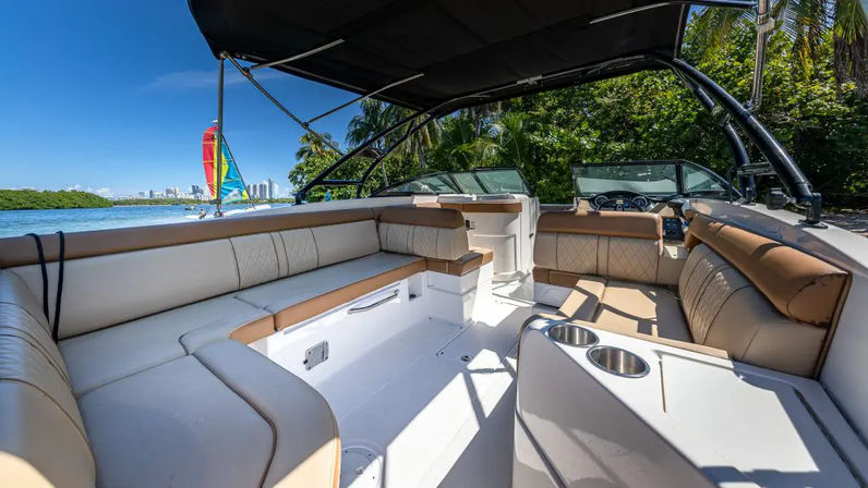 Spacious luxury motorboat interior with tan-and-white cushioned seating, stainless cup holders and black bimini top; tropical shoreline with palm trees, a colorful sailboat and distant coastal skyline under a bright blue sky.