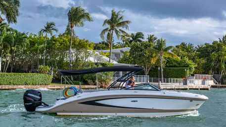 Speedboat gliding along a tropical waterfront canal lined with palm trees and private docks, two people relaxing under a black bimini top beneath a dramatic cloudy sky