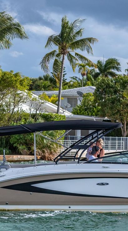 Speedboat gliding along a tropical waterfront canal lined with palm trees and private docks, two people relaxing under a black bimini top beneath a dramatic cloudy sky