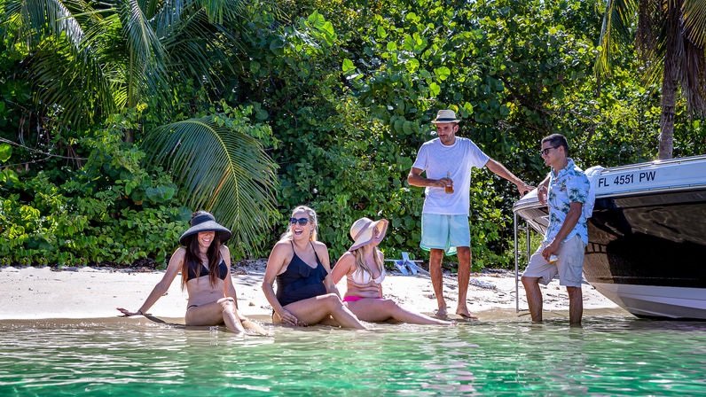 Five friends relaxing on a tropical Florida beach beside a moored boat, laughing in swimsuits and sun hats under palm trees with clear green shallow water.