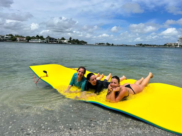 Three friends laughing on a bright yellow floating water mat in shallow bay water with waterfront homes and a blue sky dotted with clouds.