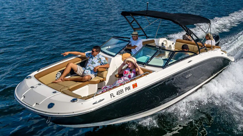 Leisure motorboat cruising through blue coastal waters on a sunny day with four adults—man reclining and pointing at bow, woman in a sunhat seated, driver at the helm and a wake trailing behind.
