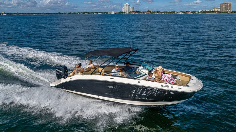 Black-and-white speedboat cruising on blue coastal waters near a city skyline, passengers relaxing under a bimini top as it cuts through the water leaving a foamy wake.