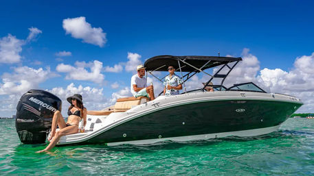 Three people relaxing on a black-and-white powerboat in clear turquoise tropical water under a bright blue sky with puffy clouds; a woman in a black bikini lounges at the stern while two men chat under the boat’s canopy.