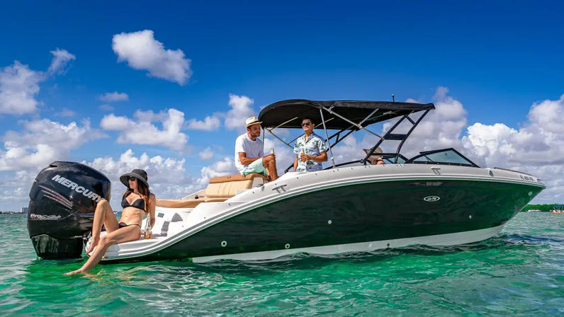 Three people relaxing on a black-and-white powerboat in clear turquoise tropical water under a bright blue sky with puffy clouds; a woman in a black bikini lounges at the stern while two men chat under the boat’s canopy.