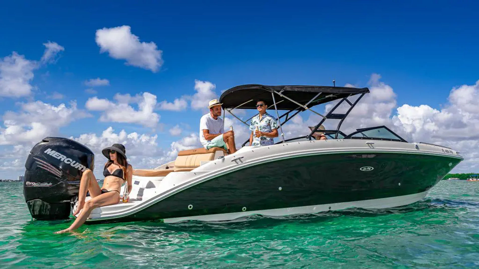 Three people relaxing on a black-and-white powerboat in clear turquoise tropical water under a bright blue sky with puffy clouds; a woman in a black bikini lounges at the stern while two men chat under the boat’s canopy.