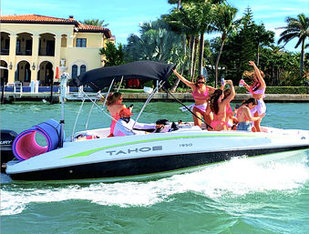 Group of friends in pink swimsuits dancing and laughing on a white motorboat with a black canopy, cruising past palm-lined waterfront and a Mediterranean‑style villa on a sunny day.