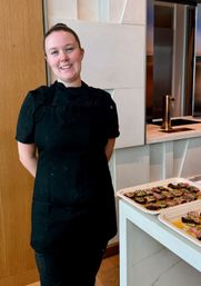 Smiling chef in black uniform standing beside trays of bite-sized plated appetizers on a white countertop in a modern kitchen