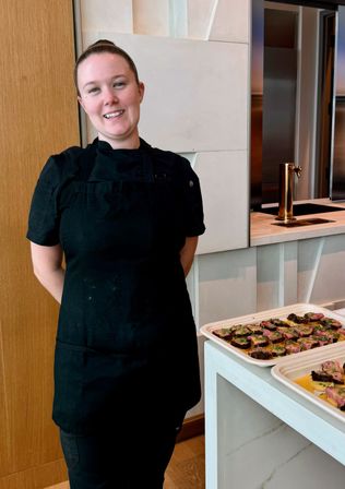 Smiling chef in black uniform standing beside trays of bite-sized plated appetizers on a white countertop in a modern kitchen