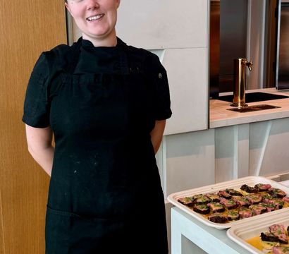 Smiling chef in black uniform standing beside trays of bite-sized plated appetizers on a white countertop in a modern kitchen