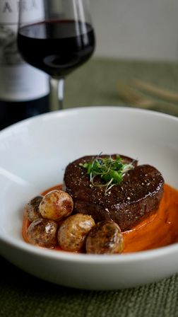 Gourmet filet mignon topped with microgreens, roasted baby potatoes on smoky red pepper sauce, plated restaurant-style with a glass of red wine
