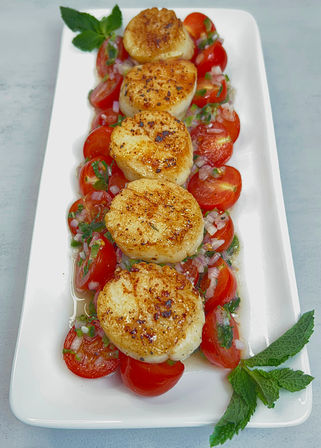 Five golden seared scallops on a white rectangular platter with cherry tomato salsa, chopped herbs and mint garnish — elegant seafood appetizer