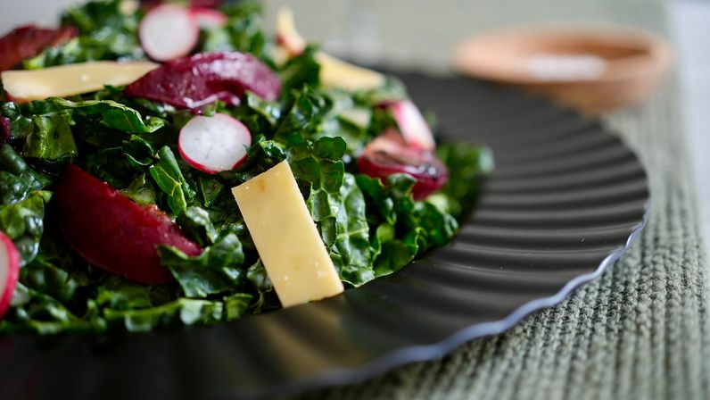 Close-up of vibrant kale salad with sliced radishes, beet wedges and shaved cheese on a black ridged plate over a textured green placemat
