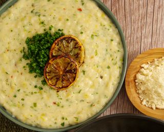 Creamy lemon-chive risotto in a green bowl, topped with charred lemon slices and a pile of chopped chives, next to a wooden bowl of grated cheese on a rustic wood table.