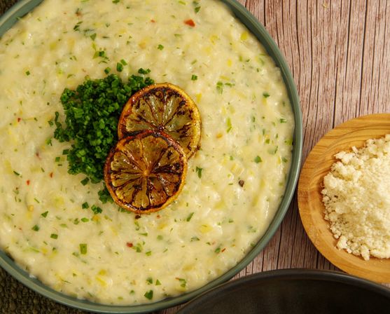 Creamy lemon-chive risotto in a green bowl, topped with charred lemon slices and a pile of chopped chives, next to a wooden bowl of grated cheese on a rustic wood table.
