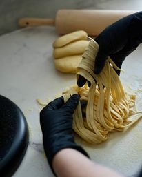 Black-gloved hands gathering fresh homemade fettuccine on a floured marble countertop with rolled dough and wooden rolling pin in the background