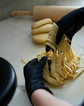 Black-gloved hands gathering fresh homemade fettuccine on a floured marble countertop with rolled dough and wooden rolling pin in the background