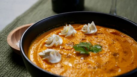 Creamy roasted pumpkin soup in a black bowl, topped with sour cream rosettes and a cilantro leaf on a green placemat — cozy autumn comfort food.