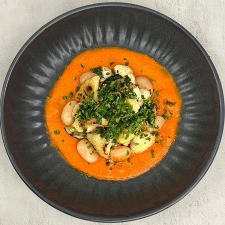 Top-down view of roasted cauliflower florets and large butter beans on bright roasted red pepper sauce, topped with chopped parsley and served in a matte black bowl on a light concrete surface.