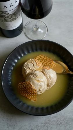 Three scoops of caramel ice cream topped with honeycomb sugar crisps in a pool of caramel sauce in a dark bowl, with a gold spoon and a glass of red wine on a gray tabletop.