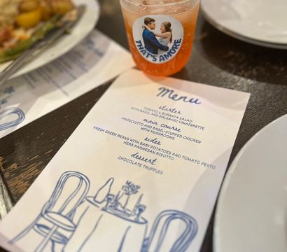 Cocktail in a plastic cup with a playful "That’s Amore" sticker beside a printed Italian-style dinner menu on a dark wooden restaurant table, plates and a partially eaten dish blurred in the background.