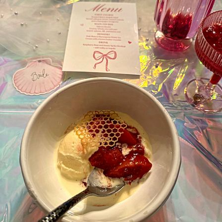 Bowl of vanilla ice cream with strawberry compote and a delicate honeycomb garnish, spoon in the bowl, set on an iridescent holographic tablecloth beside pink glassware, a bridal-themed menu card and a shell-shaped 'Bride' place tag.