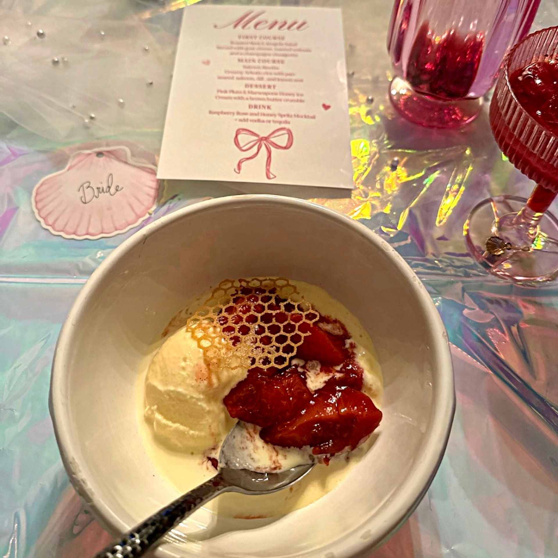 Bowl of vanilla ice cream with strawberry compote and a delicate honeycomb garnish, spoon in the bowl, set on an iridescent holographic tablecloth beside pink glassware, a bridal-themed menu card and a shell-shaped 'Bride' place tag.