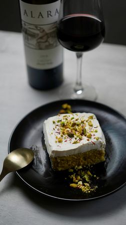 Dessert: square pistachio cake topped with whipped cream, chopped pistachios and saffron on a black plate, gold spoon beside it, red wine glass and bottle blurred in the background.
