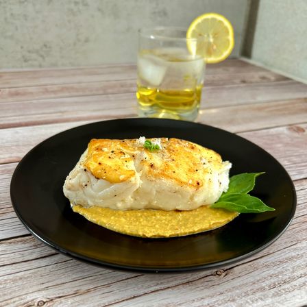 Seared cod fillet on creamy corn purée with basil garnish on a black plate, set on a rustic wooden table with a lemon-sliced cocktail in the background