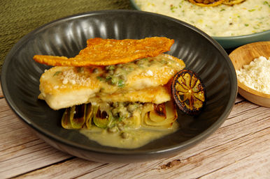 Pan-seared white fish fillets topped with caper butter, crisp cheese tuile, braised leeks and charred lemon in a black bowl on a rustic wood table — gourmet seafood entrée.