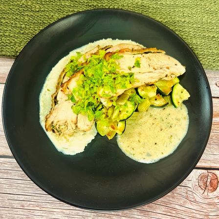 Sliced herb-roasted chicken breast over creamy mashed potatoes, sautéed zucchini and leeks with a light herb cream sauce, topped with chopped green garnish on a black plate over a rustic wooden table.