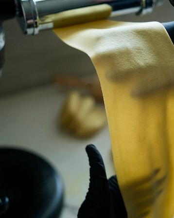 Close-up of a silky yellow sheet of homemade pasta being rolled through a stainless-steel pasta machine, a gloved hand guiding fresh pasta dough in a kitchen.