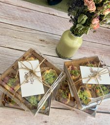 Stacked clear gift boxes of mini fruit and pistachio-topped pastries tied with twine and handwritten tags on a light rustic wooden table beside a green ceramic vase of dried pink roses
