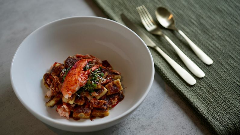 Plated lobster gnocchi in a white bowl — seared potato gnocchi topped with a bright lobster claw, brown-butter sauce and microgreens, with gold flatware on a textured green napkin beside it