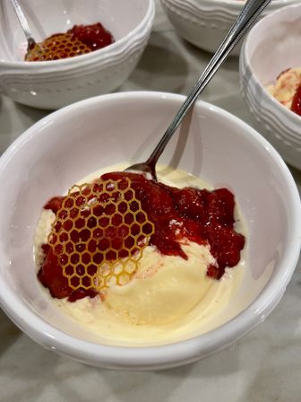 Close-up of creamy vanilla ice cream in a white ceramic bowl topped with vibrant strawberry compote and a decorative honeycomb sugar tuile, with a silver spoon.
