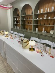 Indoor event space set for a cocktail-making workshop: long white-clothed table with mini lamps, ice buckets, shakers, martini glasses, jiggers and pink heart confetti, white folding chairs and sage-green built-in shelves with bottles and decor.