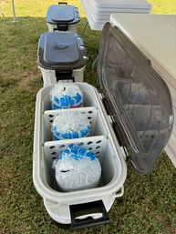 Open white portable cooler on grass at an outdoor event, showing three large clear bags of ice in separate compartments with a row of closed coolers and folding tables nearby.