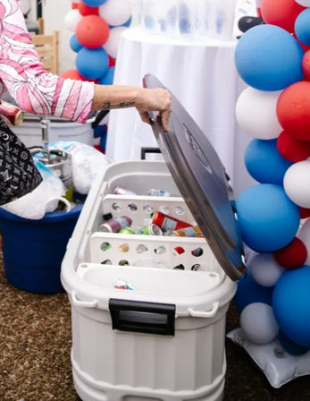 Hand lifts lid of a white cooler to reveal ice and assorted canned drinks beside red, white and blue balloon columns at an outdoor party setup.
