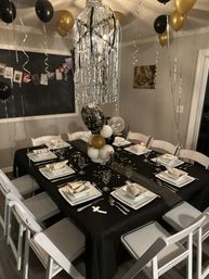 Festive black-and-gold New Year’s tablescape in a home dining room: long table with black tablecloth, white square plates and napkins, champagne glasses, balloon centerpiece and silver fringe chandelier.