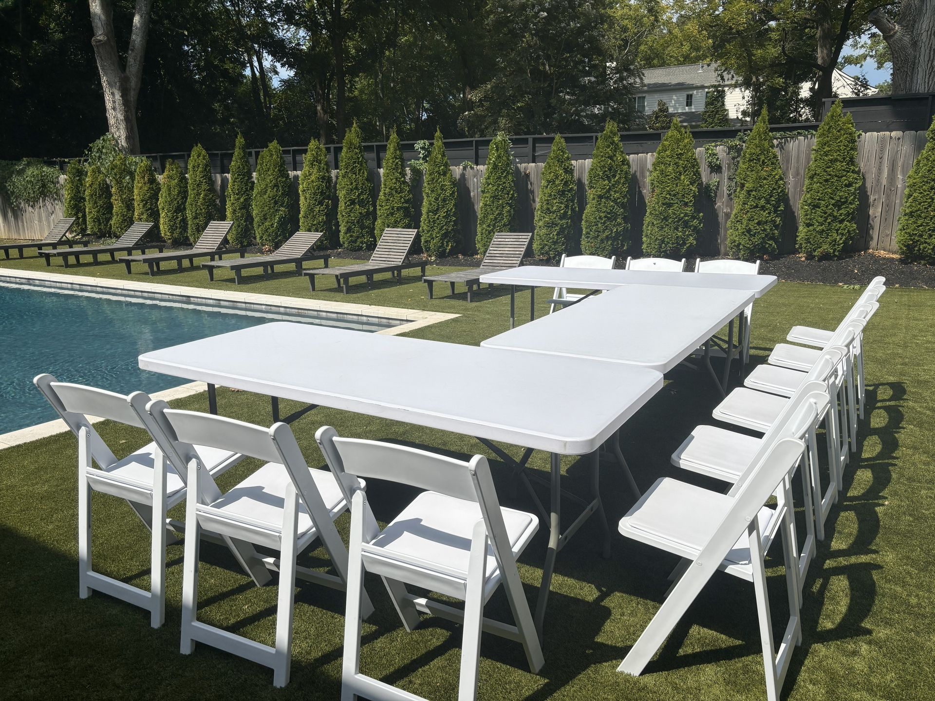 White folding tables and chairs arranged in a U-shape on green lawn by a residential swimming pool, with wooden lounge chairs and a neat row of evergreen shrubs along a privacy fence