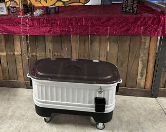 Portable rolling cooler with dark lid and white body on caster wheels in front of a wooden pallet bar draped with a pink sequin tablecloth, indoor event or market setting