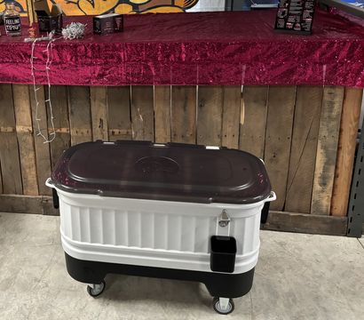 Portable rolling cooler with dark lid and white body on caster wheels in front of a wooden pallet bar draped with a pink sequin tablecloth, indoor event or market setting