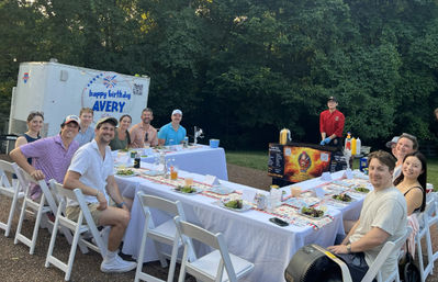 Outdoor birthday party with friends seated around a long white-tableclothed dining table, plated salads and drinks, a food cart and trailer against a wooded backyard backdrop.